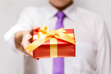 Male hand holding a gift box. Present wrapped with ribbon and bow. Christmas or birthday red package. Man in white shirt and necktie.