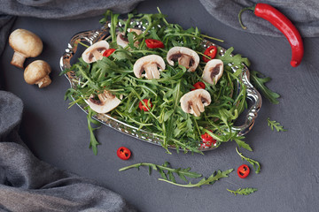 Fresh green arugula with mushrooms and chili pepper on a silver plate on dark gray background.