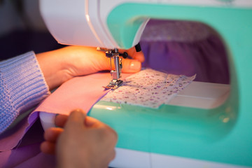 Woman`s hands in tailoring process with sewing machine of mint blue colors, lavender cotton fabrics, white tissue in lilac and pink flowers are on wooden table.