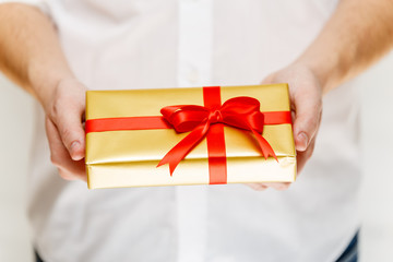 Male hands holding a gift box. Present wrapped with ribbon and bow. Christmas or birthday golden paper package. Man in white shirt.