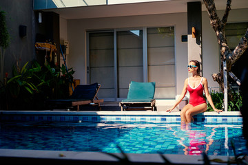 Enjoying suntan and vacation. Pretty young woman in red swimsuit sitting near swimming pool.