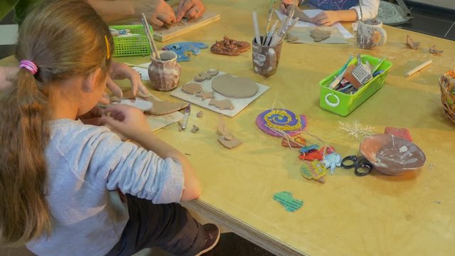 a Small Blonde Girl With a Pony Pigtail is Making Her Own Clay Toy, While a Professional Lady Helps Her in This Activity, While Sitting at a Square Table