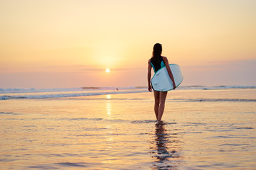 Fototapeta na wymiar Hobby and vacation. Sunset on the beach. Young woman carrying surf board.