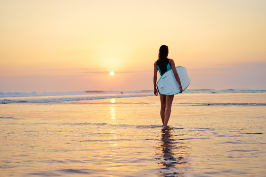 Hobby and vacation. Sunset on the beach. Young woman carrying surf board.