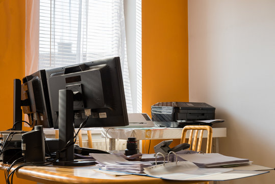 chaotic writing desk with two monitors and a printer in the background - folders, PC