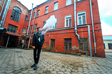 Vape. A handsome young white guy in glasses blows steam from an electronic cigarette in a vintage old red yard. Vaping.