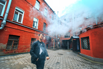 Vape. A handsome young white guy in glasses blows steam from an electronic cigarette in a vintage old red yard. Vaping.