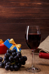 Red wine bottle. Wineglass with dark grapes branch. Gift box with bow on rustic wooden background.
