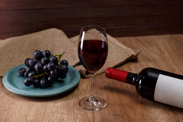 Red wine. Bottle with wineglass. Dark grapes branch on plate. On rustic wooden background.