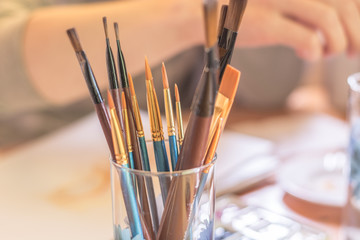 Paint brushes in glass on the table in a workshop. Master drawing at background.Close up.