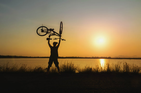 Silhouette the man stand in action lifting bicycle above his head on the meadow with sunset