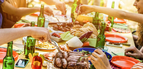 Happy friends eating barbecue meat outdoor in city park on summer time - Young people having fun drinking bear at bbq party - Friendship and food concept - Focus on sausages - Warm contrast filter