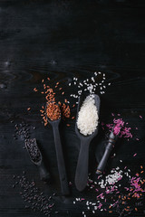 Variety assortment of raw uncooked colorful rice white, black, brown, pink in black spoons and scoop over burnt wooden background. Top view with space
