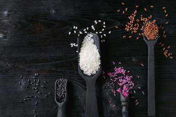 Variety assortment of raw uncooked colorful rice white, black, brown, pink in black spoons and scoop over burnt wooden background. Top view with copy space