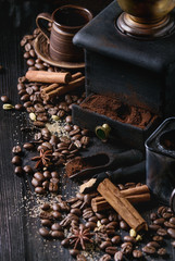 Black roasted coffee beans and grind with spices cinnamon, anise, cardamom, clove and brown sugar. With black vintage coffee grinder, scoops and terracota cup over wood burnt background.
