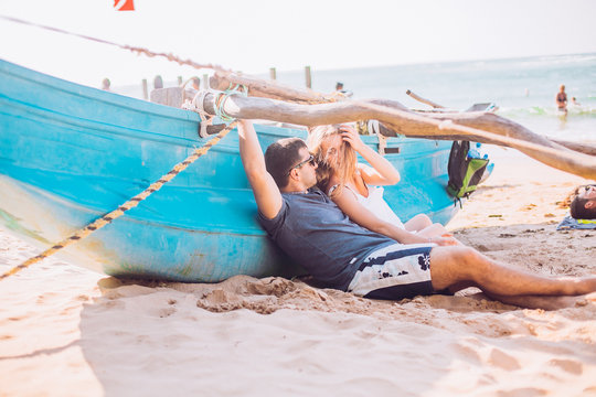 Happy Romantic Honeymoon Couple On The Beach Sitting On The Sand Near Old Fishing Boat And Looking Each Other Kissing. Husband With His Wife Near The Ocean. Sri Lanka. Hikkaduwa.