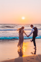 Honeymoon couple romantic in love at beach sunset. Newlywed happy young couple holding hands enjoying ocean sunset during travel holidays vacation getaway.