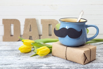 Blue cup with a mustache, gift box, yellow tulips and an inscription Dad in the background.