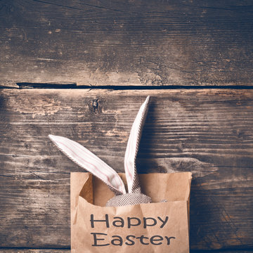 Easter bunny in a paper bag on old boards. Rabbit. Old board background. Easter ideas. Easter eggs. Space for text. Image in trendy toning. On the package text Happy Easter