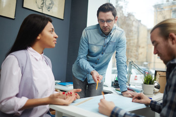 Confident young man pointing at paper with sketch and discussing it with co-workers
