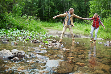 Young couple of tourists crossing river during forest adventure