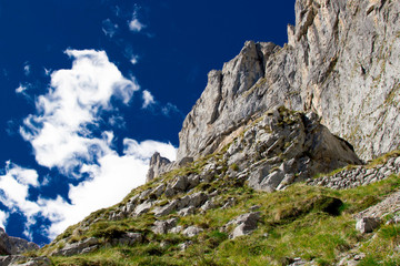 Grey rock, mountains on daytime with green grass against bright blue sky with white clouds going up - Powered by Adobe