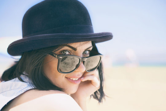 Portrait of smiling beautiful young woman in black hat and retro sunglasses on the beach