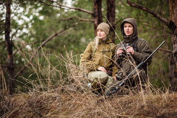 two hunters with binoculars ready to hunt, holding gun and walking in forest.