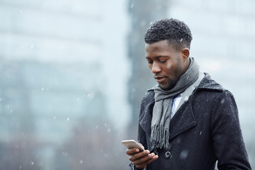 Portrait of handsome African - American businessman using smartphone phone in snowy autumn city street, typing text message and looking at screen