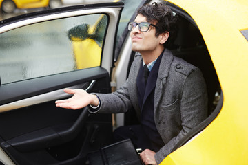 Portrait of young Middle - Eastern businessman getting out of taxi to rainy autumn street, looking displeased and testing rain with hand