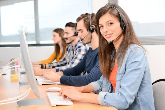 portrait of beautiful and cheerful young woman telephone operator with headset working on desktop computer in row in customer service call support helpline business center