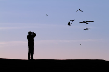 Fototapeta na wymiar Silhouette of a man on a hill taking a photographs of a small group of birds, also in silhouette, against a pale blue and orange morning sky.
