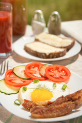 Morning breakfast: fried egg with ham, with sliced tomatoes and cucumbers, bread and butter, tomato juice in glass and bottle with newspaper on background. Cutlery, salt and pepper sunlight