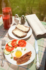 Morning breakfast: fried egg with ham, with sliced tomatoes and cucumbers, bread and butter, tomato juice in glass and bottle with newspaper on background. Cutlery, salt and pepper sunlight