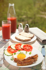 Morning breakfast: fried egg with ham, with sliced tomatoes and cucumbers, bread and butter, tomato juice in glass and bottle with newspaper on background. Cutlery, salt and pepper sunlight