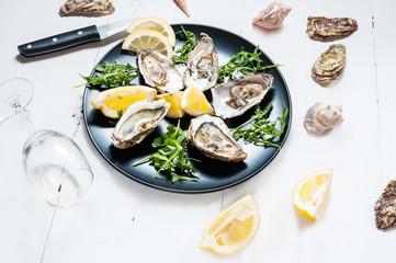 Oysters with lemon fruit on a black plate and glass of wine on a white wood table.