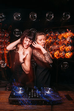 Two beautiful sexy disk jockey girls in fashionable dresses dancing and enjoying party at DJ console. Fun, nightclub, relax, light long exposure