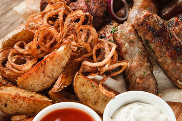 Assorted delicious grilled meat with fried potato wedges, onion rings, and sauces, close up view. Junk food, bbq, picnic concept