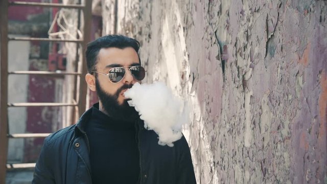 Stylish bearded man in sunglasses smokes an e-cigarette on a red wall background