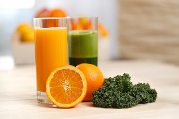 Orange juice and green vegetable juice in tall glasses with orange half kale and fruit basket in the background