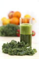 Green vegetable juice in a tall glass with a leaf of kale and a basket of fruits and vegetables in the background