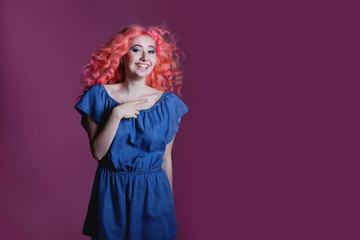 Beautiful girl with pink hair in blue dress shows sign on lilac background, place for text