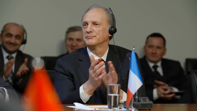 French delegate at international summit or forum. Middle aged caucasian polititian man in suit applauds someone's speech at meeting. He listen the speaker, nods approvingly and claps his hands.