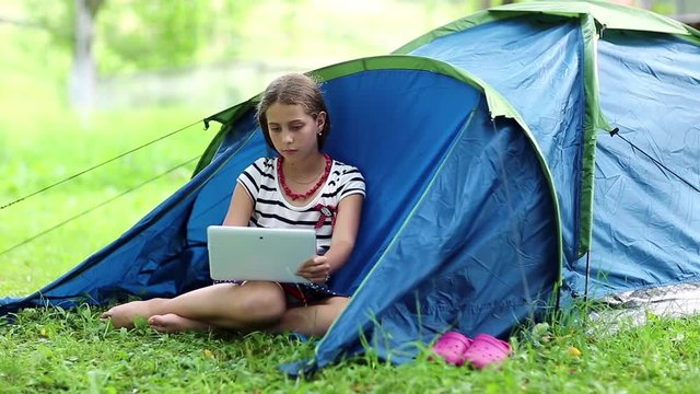 Girl sits on green grass near tourist tent and uses tablet computer. Girl looks on display of tablet pc. Girl with tablet pc sits on green grass near tourist tent