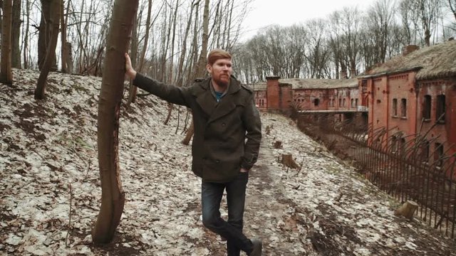 A young man With a beard, in a gray coat walks through the park in the spring or autumn trees yellow leaves, next to an abandoned German fort, brick walls, an old iron fence