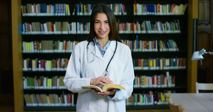 A young and beautiful female doctor in a library smiling happy and holding books after doing a search and after studying. Concept: educational, portrait, library, and medical care and welfare