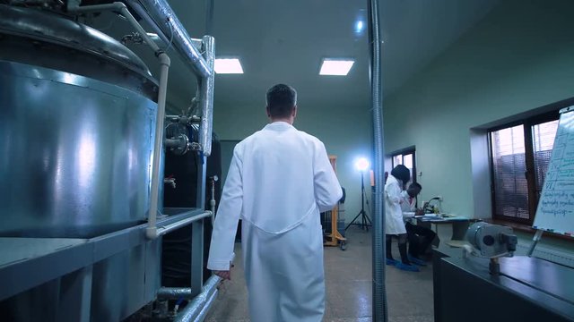 Rear view video of unrecognizable male scientist in white uniform walking through lad with metal tubes and engineering equipment aside