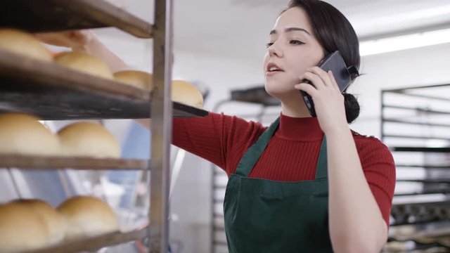 Worker in bakery kitchen talking on phone as she checks trays of bread