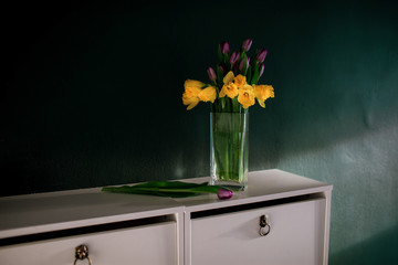 yellow daffodil flowers with purple tulip blooming in vase with green wall next wicked basket