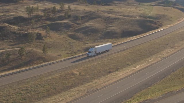 AERIAL Flying above freight container semi truck driving along the empty highway running through sunny bumpy hilly landscape. Trailer transporting goods across United States traveling on interstate
